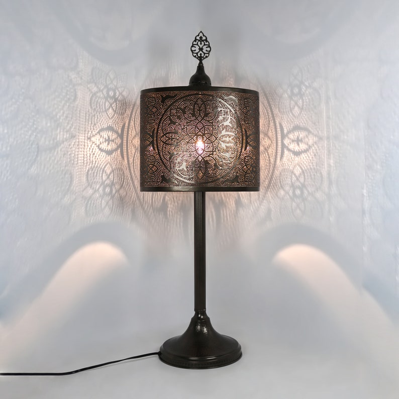 K&ouml;nnte beinhalten: Eine schwarze Tischlampe mit einem dekorativen Metallschirm mit geometrischem Muster. Die Lampe hat einen hohen, schlanken Sockel und einen Abschluss oben.