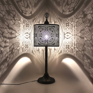 Peut inclure: Une lampe de table en métal noir avec un abat-jour carré à motifs complexes. L'abat-jour projette une ombre décorative sur le mur derrière elle.