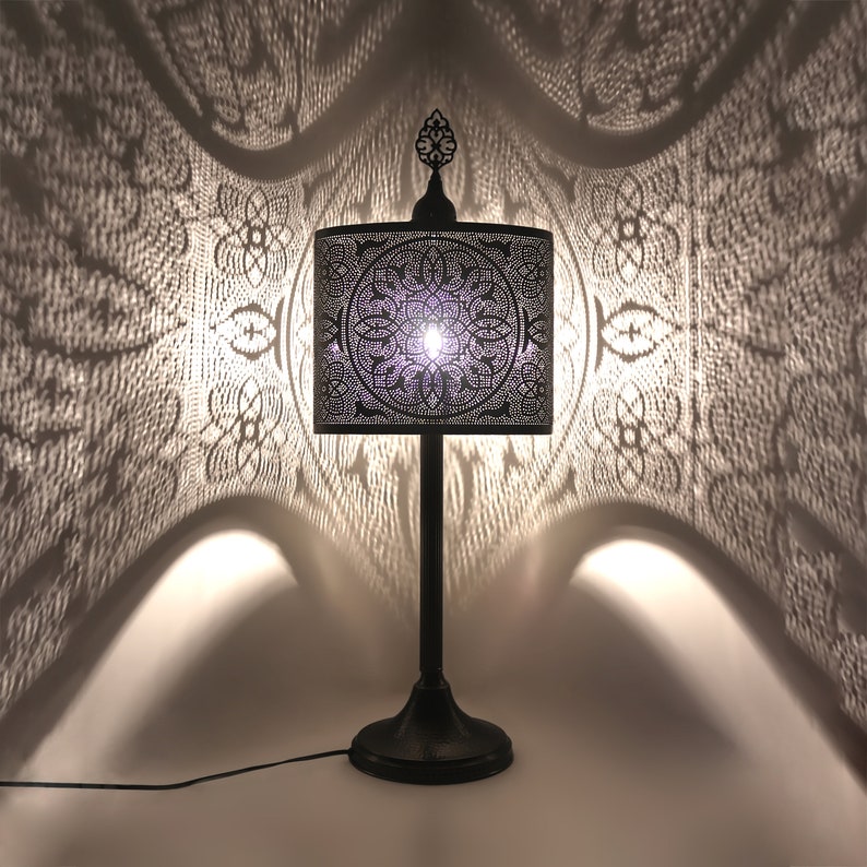 K&ouml;nnte beinhalten: Eine schwarze Tischlampe aus Metall mit einem quadratischen, filigran verzierten Lampenschirm. Der Lampenschirm wirft einen dekorativen Schatten auf die Wand dahinter.