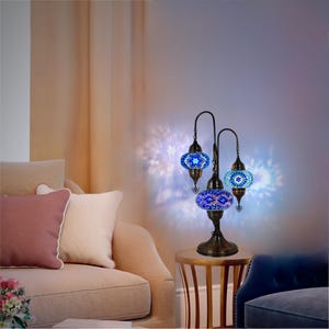 May include: A three-light table lamp with a bronze base and three blue mosaic glass shades. The lamp is on a wooden end table with a blue sofa in the background.