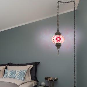 K&ouml;nnte beinhalten: Eine H&auml;ngeleuchte mit einem roten Sternmuster auf wei&szlig;em Grund. Die Lampe hat einen Metallrahmen und eine Kette. Die Lampe h&auml;ngt in einem Schlafzimmer mit grauer Wand.