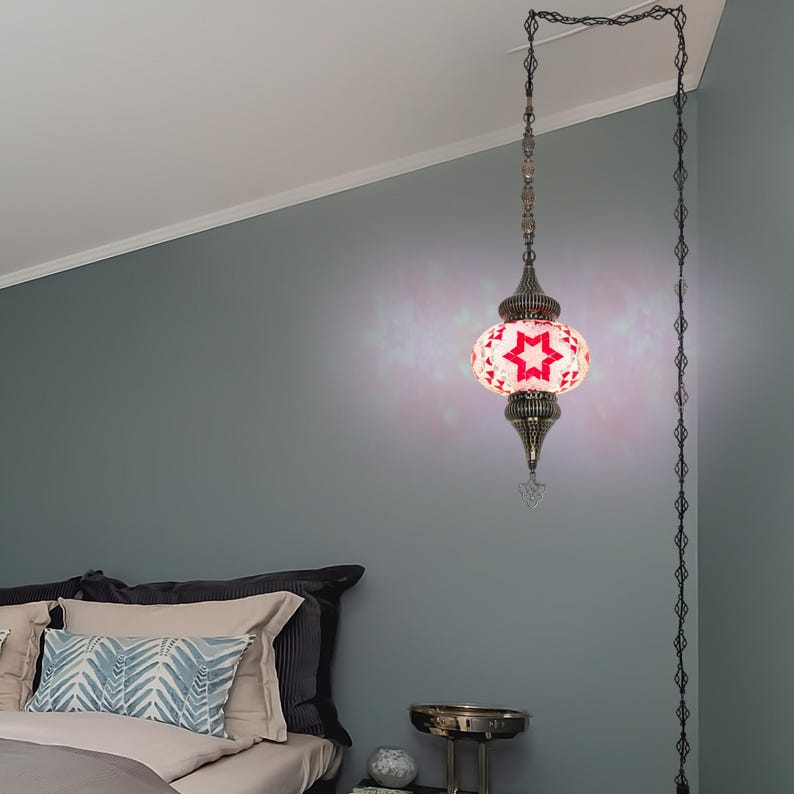 K&ouml;nnte beinhalten: Eine H&auml;ngeleuchte mit einem roten Sternmuster auf wei&szlig;em Grund. Die Lampe hat einen Metallrahmen und eine Kette. Die Lampe h&auml;ngt in einem Schlafzimmer mit grauer Wand.