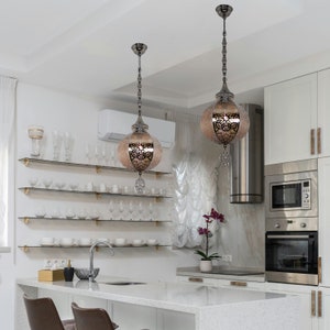 Peut inclure: Deux luminaires suspendus ornés, avec des ouvrages en métal complexes et un globe en verre dépoli. Les luminaires sont suspendus à un plafond blanc dans une cuisine moderne.