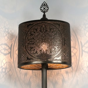K&ouml;nnte beinhalten: Eine schwarze Metall-Stehlampe mit einem dekorativen, verzierten Abschluss und einem runden, perforierten Lampenschirm. Der Lampenschirm hat ein goldenes Innenleben und ein Muster aus filigranen Blumenmotiven.