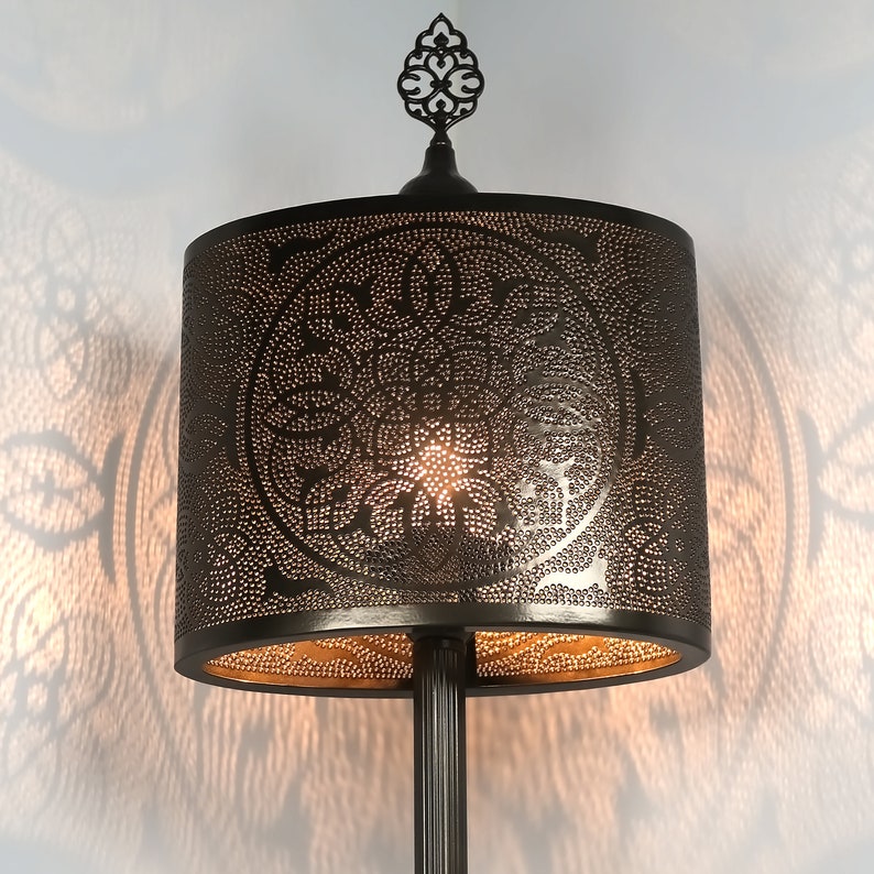 K&ouml;nnte beinhalten: Eine schwarze Metall-Stehlampe mit einem dekorativen, verzierten Abschluss und einem runden, perforierten Lampenschirm. Der Lampenschirm hat ein goldenes Innenleben und ein Muster aus filigranen Blumenmotiven.