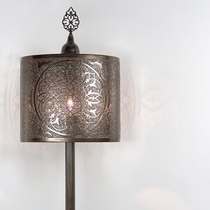 K&ouml;nnte beinhalten: Eine bronzene Stehlampe mit einem zylindrischen Schirm, der mit einem filigranen, komplizierten Ausschnittmuster verziert ist. Der Schirm ist mit einem dekorativen Abschlussst&uuml;ck versehen.