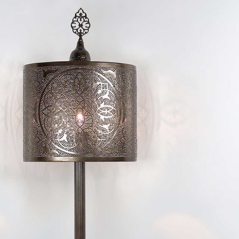 K&ouml;nnte beinhalten: Eine bronzene Stehlampe mit einem zylindrischen Schirm, der mit einem filigranen, komplizierten Ausschnittmuster verziert ist. Der Schirm ist mit einem dekorativen Abschlussst&uuml;ck versehen.