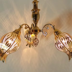 May include: A gold-toned chandelier with three ornate, openwork tulip-shaped shades. The chandelier has a decorative, floral design.