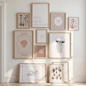 May include: A collection of framed art prints with a coastal theme. The prints feature illustrations of seashells, a jellyfish, and a mermaid, along with the text "Let's all be Mermaids" and "Stay wild ocean child". The frames are light wood.