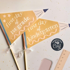 Op de afbeelding: Twee gele vlaggen met een potloodontwerp. De bovenste vlag zegt "eerste dag van de 1e klas" en de onderste vlag zegt "eerste dag van de kleuterschool".