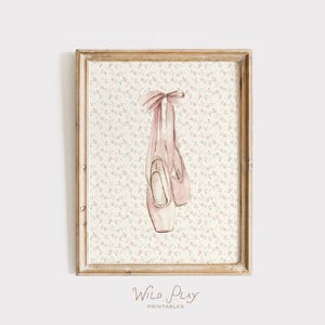 May include: A framed print featuring a watercolor illustration of pink ballet pointe shoes with ribbons. The background has a floral pattern. The print is in a wooden frame. The text "Wild Play Printables" is at the bottom.