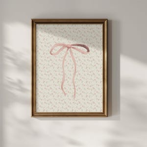 May include: A framed print with a watercolour illustration of a pink bow. The bow is central on a background of small pink flowers and green leaves on a cream backdrop. The frame is a warm, golden brown.