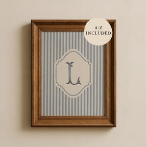 Puede incluir: Una impresión enmarcada con un marco de madera marrón claro. La impresión tiene un fondo a rayas en tonos grises y blancos. Un emblema color crema en el centro muestra la letra "L" en color gris oscuro. El texto "A-Z INCLUDED" está en un círculo color crema.