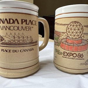 Puede incluir: Dos tazas de cerámica beige con bordes blancos. La taza izquierda presenta el texto Canada Place Vancouver y Place du Canada con un dibujo lineal del edificio. La derecha muestra un gráfico de la cúpula de la Expo 86 y Vancouver Canada.