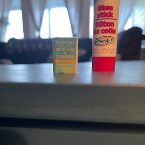 May include: A small blue and yellow Webster's All-In-One Dictionary & Thesaurus book next to a red glue stick with the brand name Rose Art and the text "Glue stick Baton de colle" on the label.