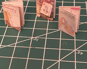 May include: Three folded paperbacks on a green cutting mat. The books are titled "Embroidery Basics", "Somerset Studio", and "Embroidered Country Gardens".