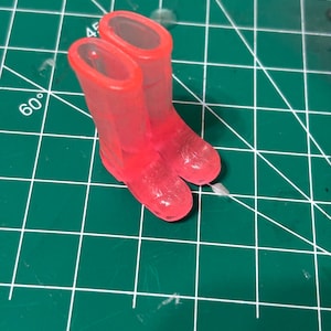 May include: A pair of miniature, translucent red rain boots. The boots are small, with a rounded toe and a short shaft. They are made of a clear, glossy material, and are set against a green cutting mat.