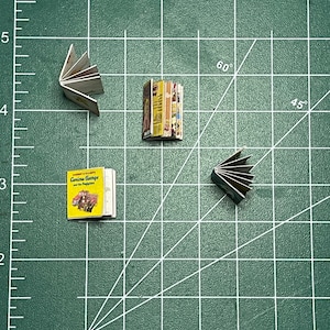 May include: Three miniature books on a green cutting mat. The books are all different sizes and have different covers. One book is yellow with a picture of a car on the cover and the text "Curious George and the Firemen". Another book is a stack of colorful books with a white spine. The third book is a stack of white books with a black spine.