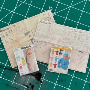 Op de afbeelding: Drie Simplicity-naaipatronen voor babykleding. De patronen liggen op een groene snijmat. De patronen zijn voor een baby-wikkeldeken, een baby-slaapzak en een baby-muts. De patronen zijn allemaal in dezelfde stijl en hebben een witte achtergrond met zwarte tekst.