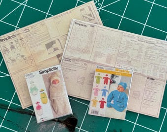 May include: Three Simplicity sewing patterns for baby clothes. The patterns are on a green cutting mat. The patterns are for a baby bunting, a baby sleeper, and a baby hat. The patterns are all in the same style and have a white background with black text.