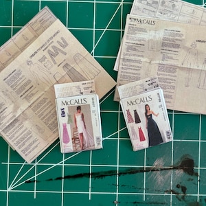 May include: Two McCall's sewing patterns for dresses. The patterns are folded and show the front and back of the dresses. The patterns are on a green cutting mat.