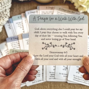 May include: A card with the text "A Prayer for a Walk With God" is held in front of a collection of similar cards. The card features a prayer from Deuteronomy 6:5, with floral accents. The background includes a soft, neutral-toned fabric and floral elements.