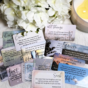 May include: A collection of inspirational cards with various phrases and Bible verses, such as "Love," "Power," and "Seek." The cards feature soft pastel backgrounds and elegant typography. White flowers and a lit candle are in the background.