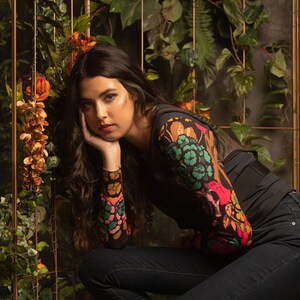 May include: A woman wearing a black top with a colorful floral print and dark blue jeans sits on a stool in front of a green leafy backdrop.