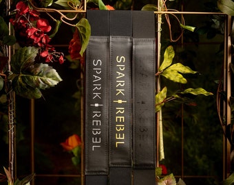 May include: Three black elastic straps with gold lettering that reads "SPARK REBEL". The straps have silver metal snaps.