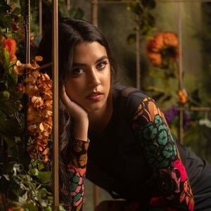 May include: A woman with long dark hair is wearing a black long-sleeved shirt with a colorful floral print. She is sitting with her chin resting on her hand and looking at the camera.