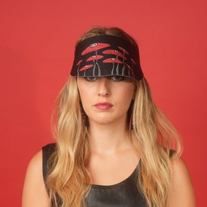 Puede incluir: Una visera de cuero negro con un diseño de amapolas rojas. La visera la lleva puesta una persona con el pelo largo y rubio.