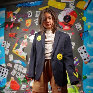 May include: A young person wearing a navy blue blazer with yellow smiley face patches, a white floral print shirt, and brown cargo pants. The person is standing in front of a colorful wall with various images and patterns.
