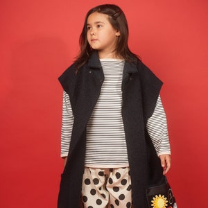 May include: A young person wearing a black sleeveless jacket over a white and black striped long-sleeved shirt. They are also wearing black and white polka dot pants and a black bag with colorful patches.