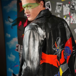 May include: A black leather jacket with a painted design of a robot on the back. The robot is red, blue, and yellow and has a red cape. The jacket has a zipper closure and a collar.