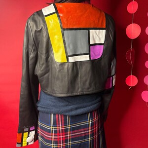 May include: Black leather jacket with a colorful geometric design on the back and sleeves. The design includes orange, yellow, gray, white, and purple blocks. The jacket is cropped and paired with a blue sweater and a plaid skirt.