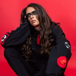 May include: A person wearing a black hoodie with red and white patches, black pants, and black shoes. The person has long dark hair and black makeup around their eyes.
