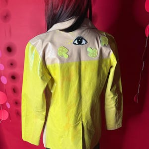 May include: A yellow jacket with a tan collar and shoulder area. The back of the jacket features an eye and two mushroom-shaped designs. The jacket has a glossy finish and a split in the back.