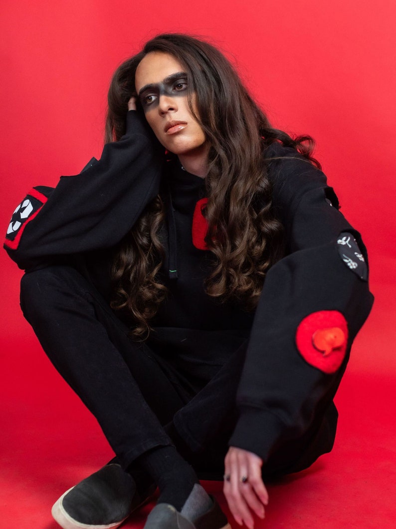 May include: A person wearing a black hoodie with red and white patches, black pants, and black shoes. The person has long dark hair and black makeup around their eyes.