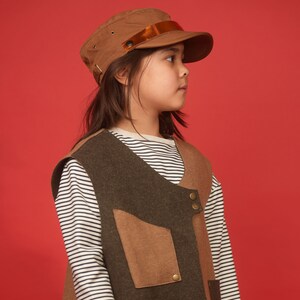 May include: A young person wearing a brown and green vest with a brown cap and a white and black striped long-sleeved shirt.