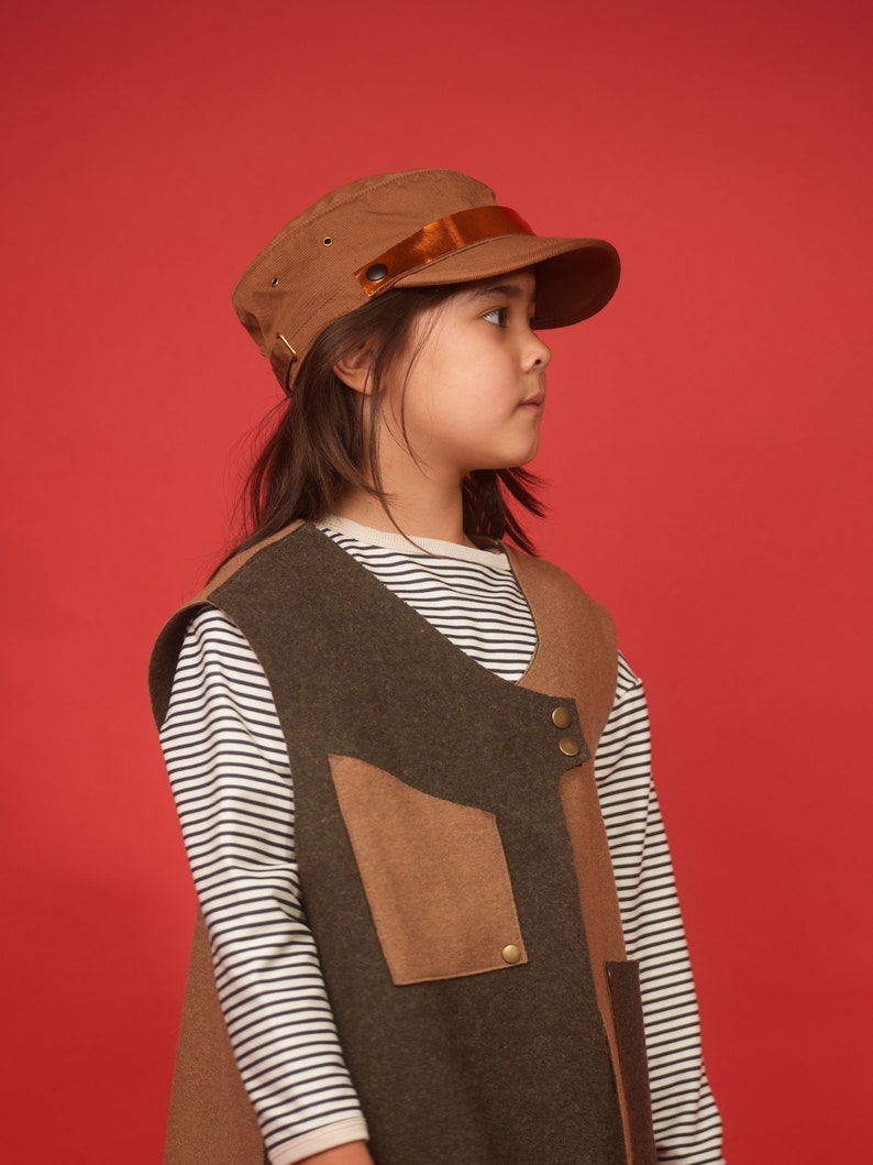 May include: A young person wearing a brown and green vest with a brown cap and a white and black striped long-sleeved shirt.