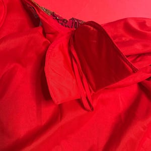 May include: A vibrant red garment with a flowing, silky texture. The neckline is adorned with gold trim and dark embellishments. The fabric is gathered and draped, creating soft folds and shadows.