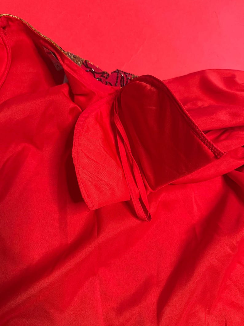 May include: A vibrant red garment with a flowing, silky texture. The neckline is adorned with gold trim and dark embellishments. The fabric is gathered and draped, creating soft folds and shadows.