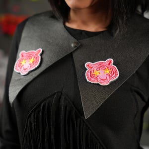May include: Black top with a leather collar featuring two embroidered tiger patches. The patches are pink with yellow star-shaped eyes. The top has a fringe detail.