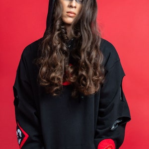 May include: A person with long, curly brown hair is wearing a black hooded sweatshirt with red accents and a red patch with a speech bubble on the sleeve. The person has black makeup around their eyes.