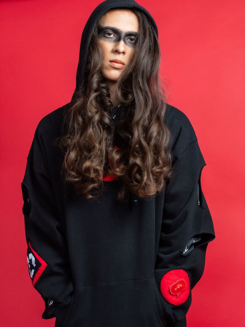 May include: A person with long, curly brown hair is wearing a black hooded sweatshirt with red accents and a red patch with a speech bubble on the sleeve. The person has black makeup around their eyes.