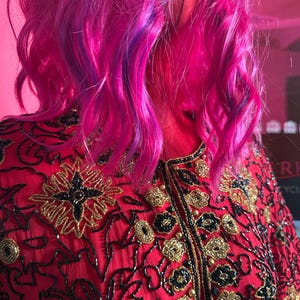 May include: A red jacket with intricate gold and black embroidery. The jacket features a high collar and is adorned with floral and geometric patterns. The model has vibrant pink and purple hair.