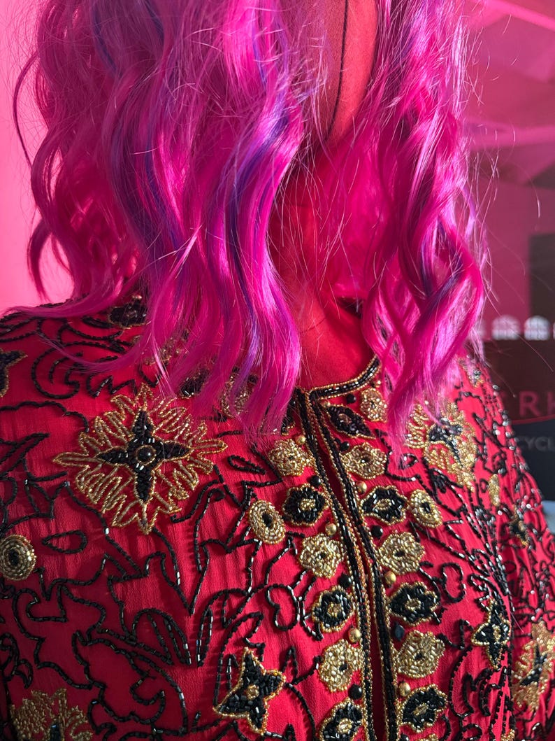 May include: A red jacket with intricate gold and black embroidery. The jacket features a high collar and is adorned with floral and geometric patterns. The model has vibrant pink and purple hair.