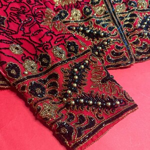May include: A red jacket with intricate beadwork in black and gold. The jacket features ornate floral and abstract patterns, with a detailed design on the sleeves. The garment is embellished with small, round beads and sequins.