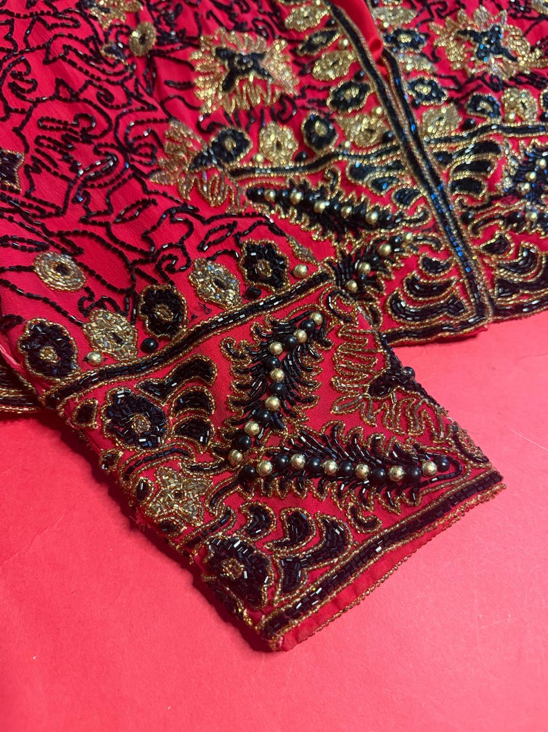 May include: A red jacket with intricate beadwork in black and gold. The jacket features ornate floral and abstract patterns, with a detailed design on the sleeves. The garment is embellished with small, round beads and sequins.