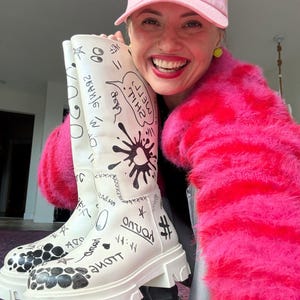 May include: A pair of white knee-high boots featuring black hand-drawn graffiti-style illustrations, including text, stars, and abstract shapes. The boots have a thick, chunky white lug sole and a smooth finish with various black ink markings.